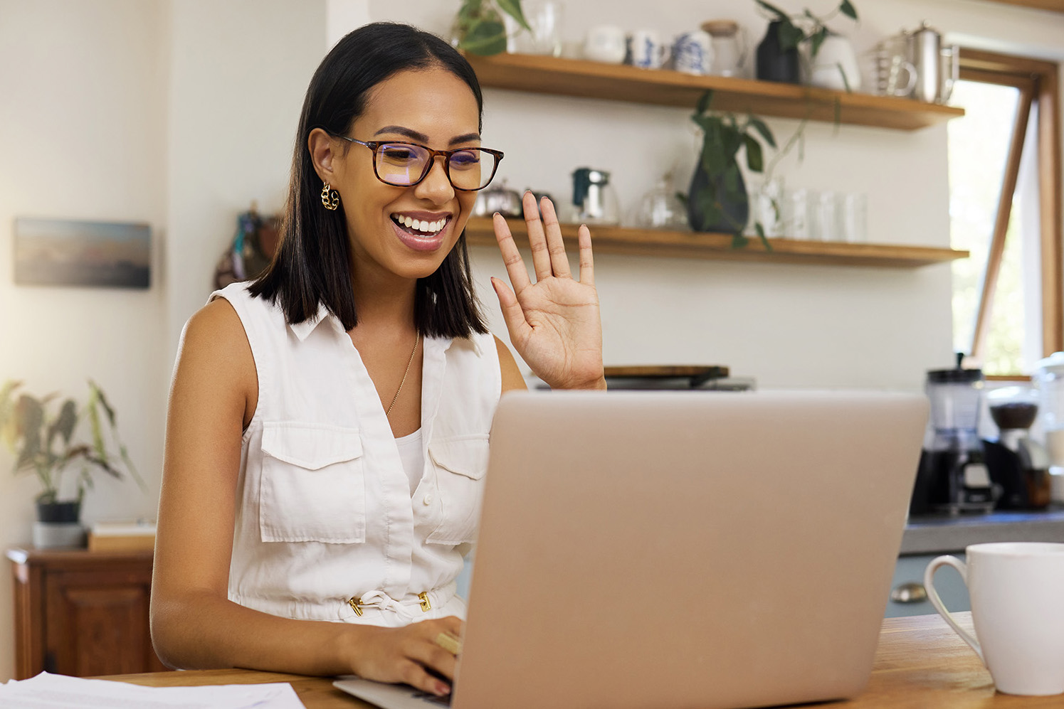 A smiling woman waving to her laptop at the start of a video call, at a table in her kitchen