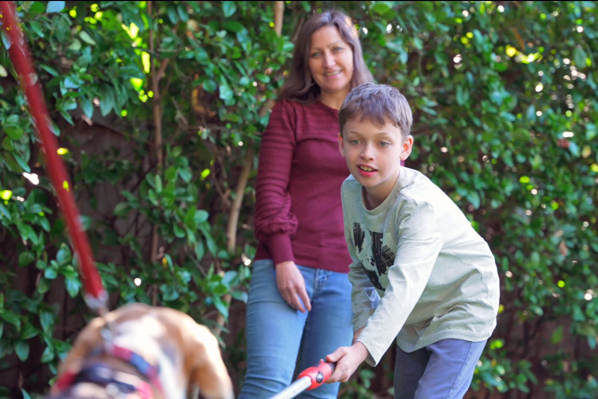 A boy guiding a dog with a target stick while his mum watches