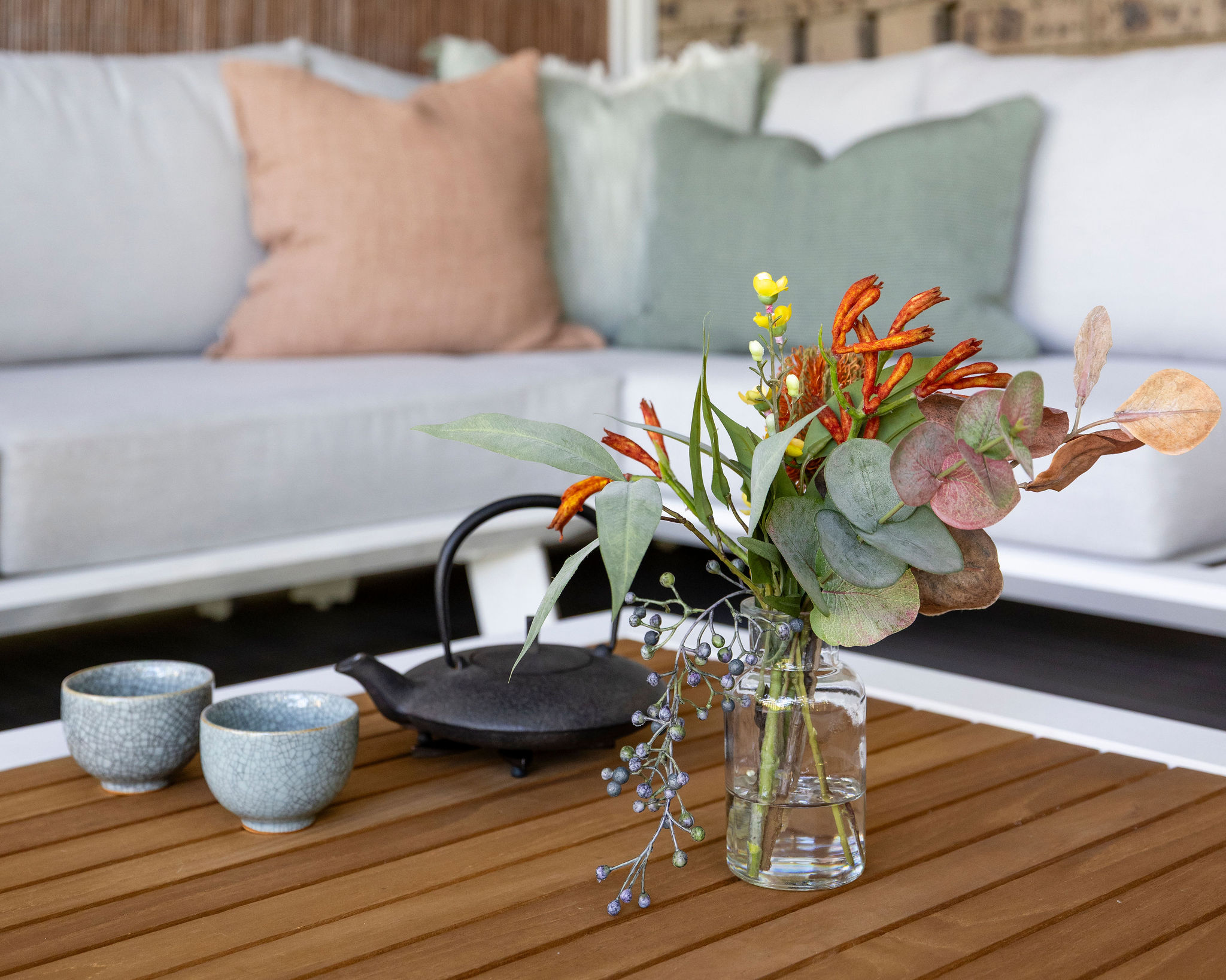 A cast-iron teapot, two cups and a native flower arrangement on the deck's timber table, with the lounge seating behind