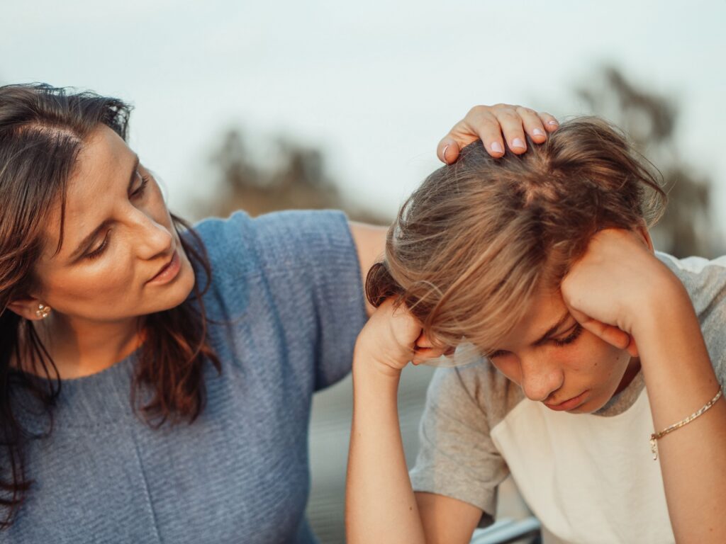 A boy holds his head in his hands while he is comforted by his mother