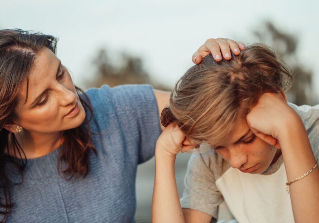 A boy holds his head in his hands while he is comforted by his mother
